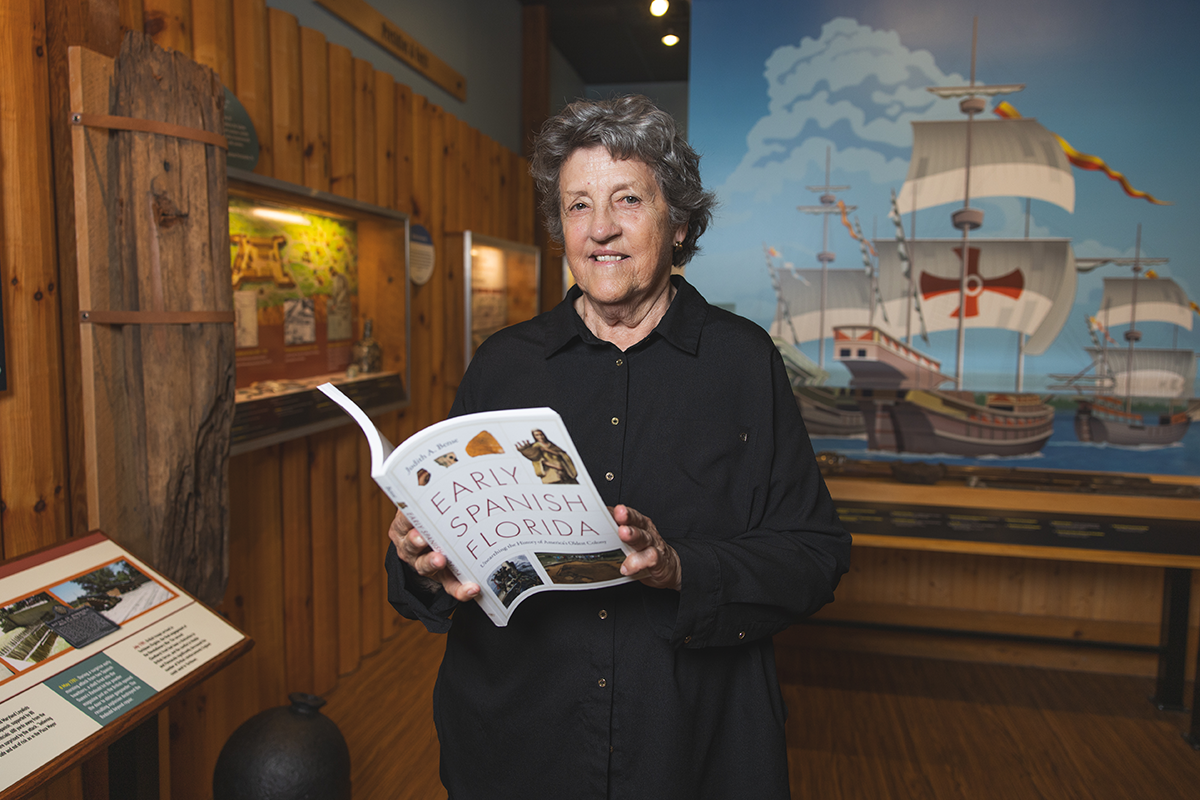 Dr. Judy Bense holds her book, "Early Spanish Florida," inside a museum with a Spanish ship painted on the wall behind her.