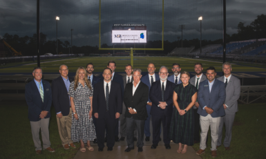 A group of fifteen people stand in front of a football field goal post with a video board in the back that reads, "Michles & Booth. Official Law Firm of UWF Athletics."