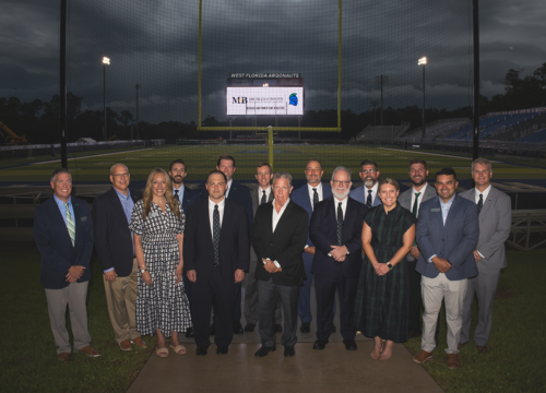 A group of fifteen people stand in front of a football field goal post with a video board in the back that reads, "Michles & Booth. Official Law Firm of UWF Athletics."