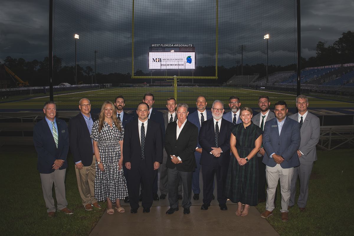 A group of fifteen people stand in front of a football field goal post with a video board in the back that reads, "Michles & Booth. Official Law Firm of UWF Athletics."