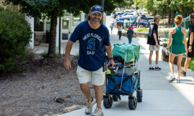 Father wearing West Florida Dad t-shirt pulls a wagon on a sidewalk leading into a dorm.