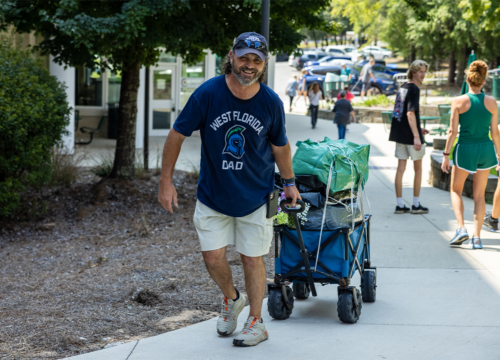Father wearing West Florida Dad t-shirt pulls a wagon on a sidewalk leading into a dorm.