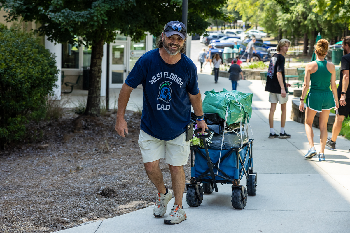 Father wearing West Florida Dad t-shirt pulls a wagon on a sidewalk leading into a dorm.