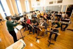 A music professor conducting a class of 16 music students seated indoors in three rows playing various instruments from saxophones, trombones, trumpets, and more.