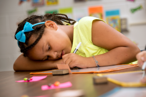 An elementary-aged student writing on paper while seated at an indoor table with their head leaned down on one arm..