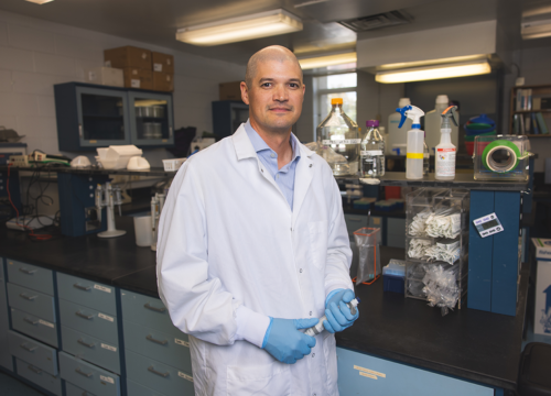 Researcher in white coat stands in science lab holding test tube.