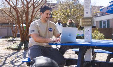 A student wearing a UWF Student Veterans of America tshirt sits at a picnic table outside the University Commons working on a laptop.