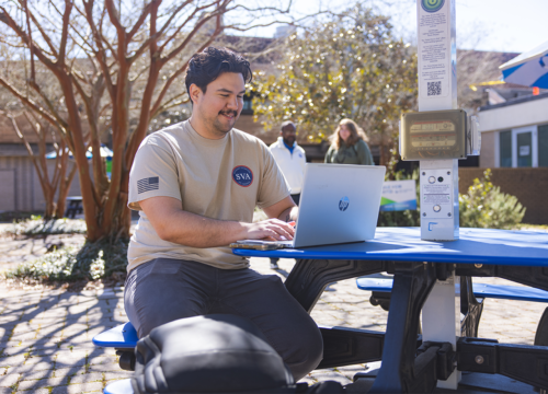 A student wearing a UWF Student Veterans of America tshirt sits at a picnic table outside the University Commons working on a laptop.