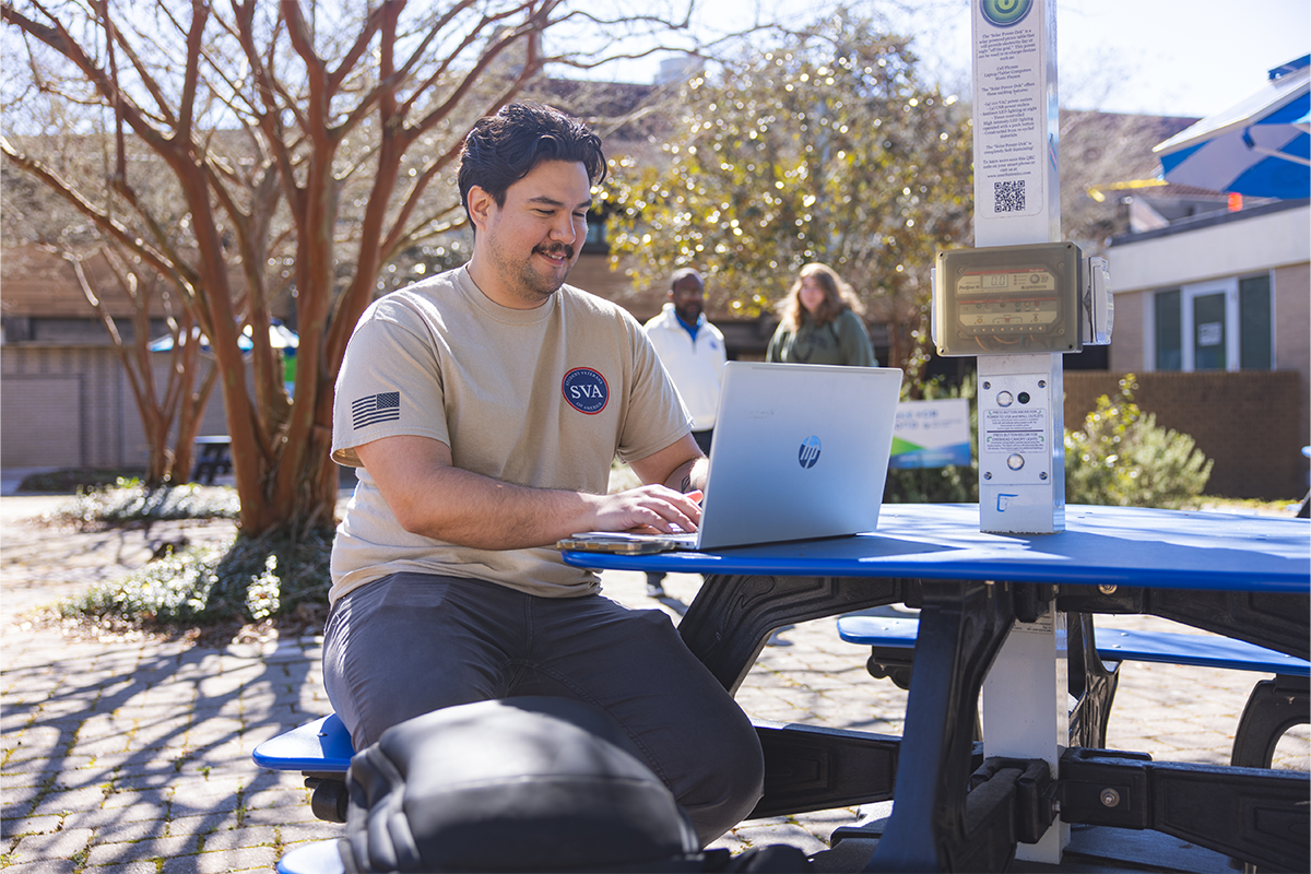 A student wearing a UWF Student Veterans of America tshirt sits at a picnic table outside the University Commons working on a laptop.