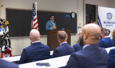 Manny Diaz Jr. speaks at a podium in front of a room of people. Banner on the right says, "UWF Office of Military Services."