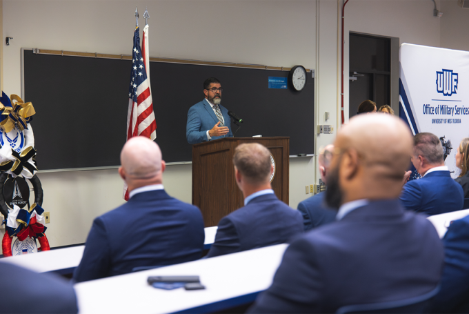 Manny Diaz Jr. speaks at a podium in front of a room of people. Banner on the right says, "UWF Office of Military Services."