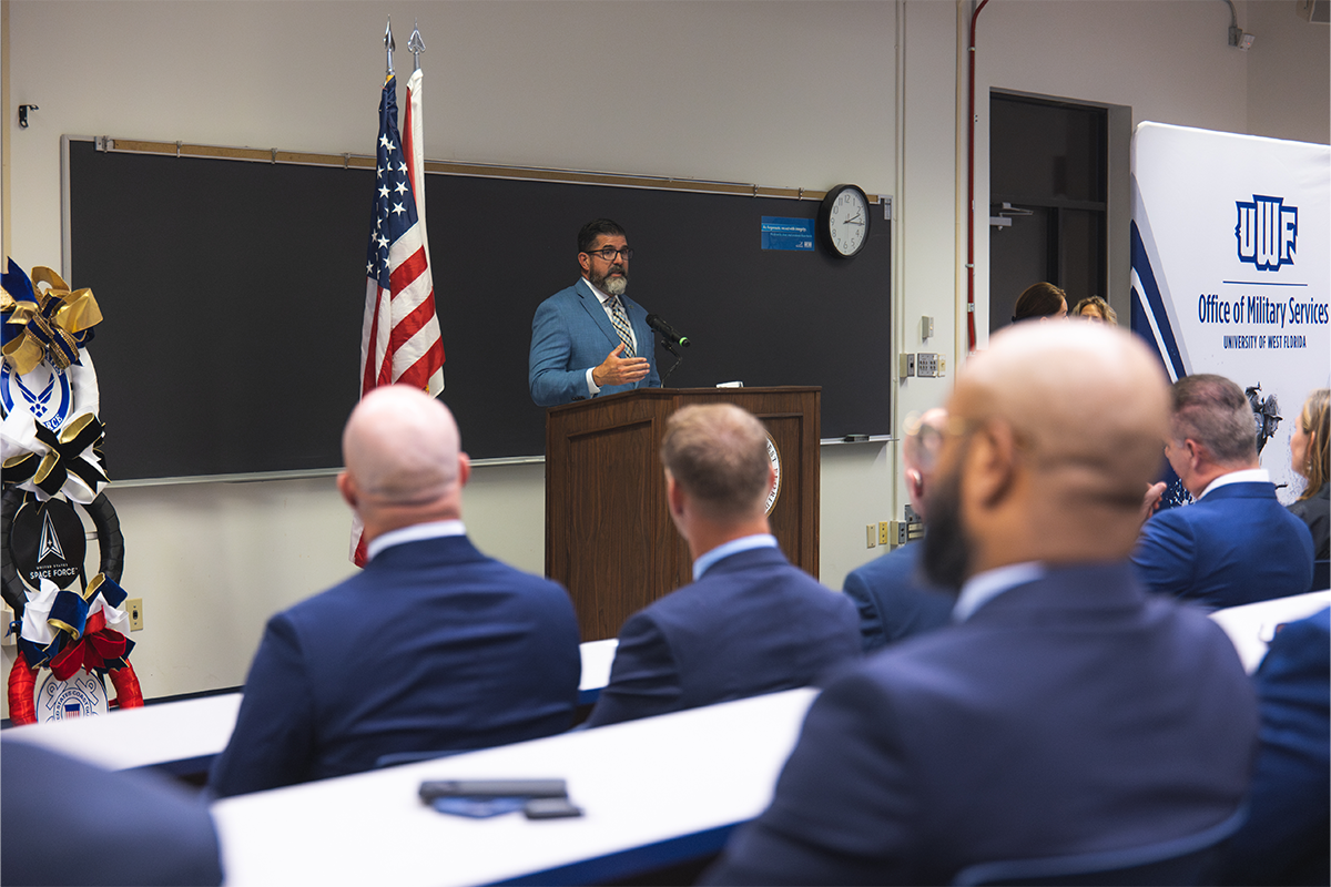 Manny Diaz Jr. speaks at a podium in front of a room of people. Banner on the right says, "UWF Office of Military Services."