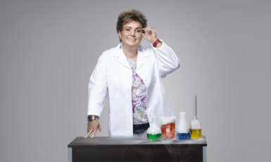 Chemistry professor stands at a desk with test tubes and wears a white coat and lab goggles.