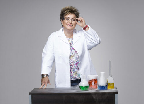 Chemistry professor stands at a desk with test tubes and wears a white coat and lab goggles.
