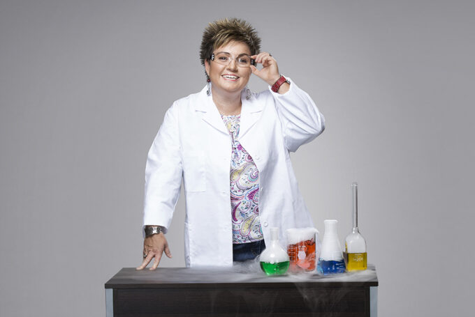 Chemistry professor stands at a desk with test tubes and wears a white coat and lab goggles.