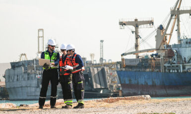 Three professional port workers stand on the gravel ground of a busy industrial port fully equipped with safety gears including hard hats and high-visibility vests while collaborating using a laptop and clipboard.