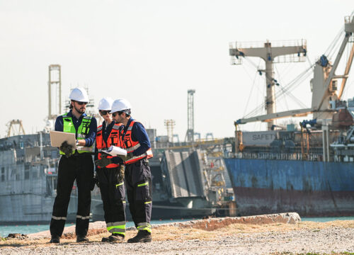 Three professional port workers stand on the gravel ground of a busy industrial port fully equipped with safety gears including hard hats and high-visibility vests while collaborating using a laptop and clipboard.