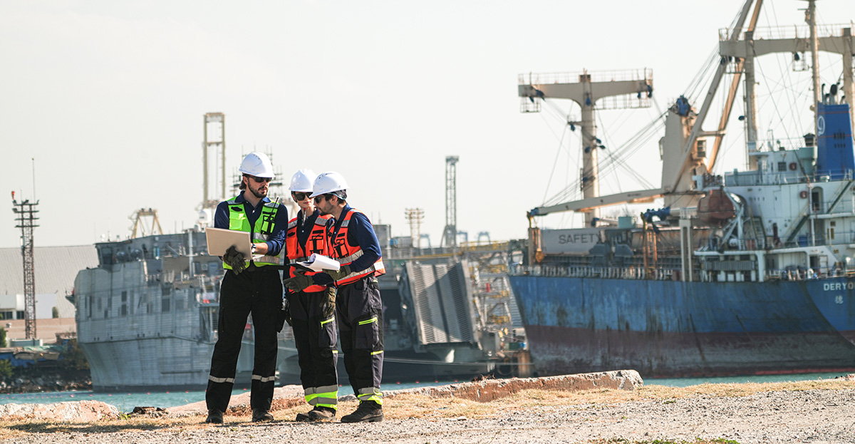 Three professional port workers stand on the gravel ground of a busy industrial port fully equipped with safety gears including hard hats and high-visibility vests while collaborating using a laptop and clipboard.