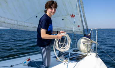 Student kneels on the bow of a sailboat with rope in hand.