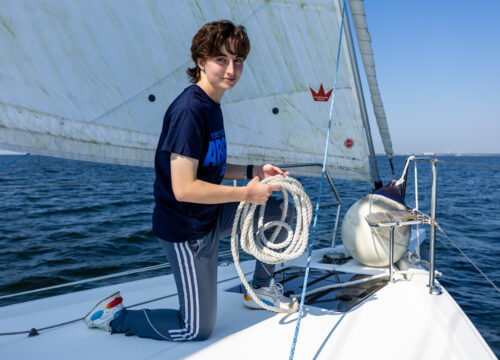 Student kneels on the bow of a sailboat with rope in hand.