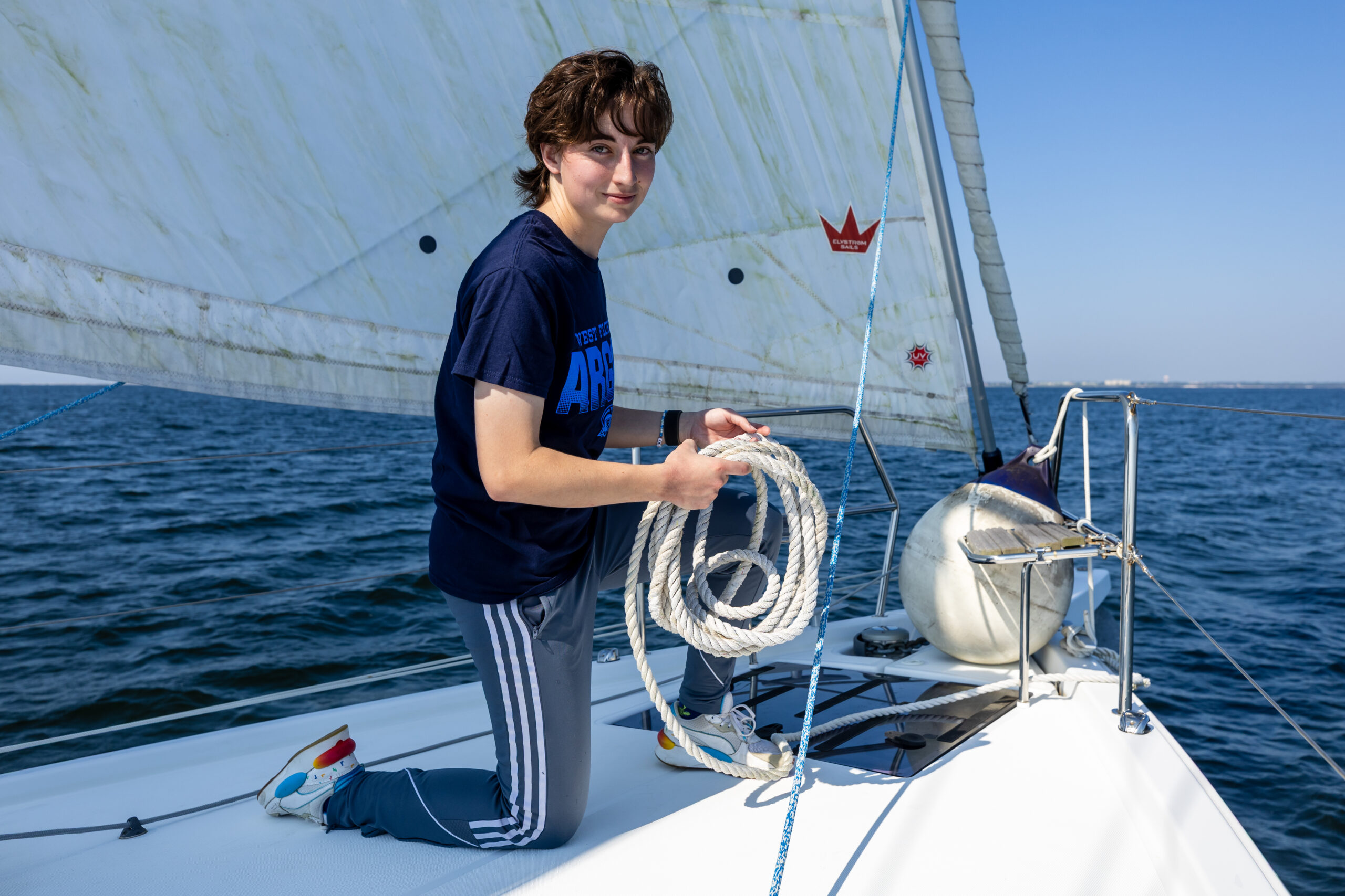 Student kneels on the bow of a sailboat with rope in hand.