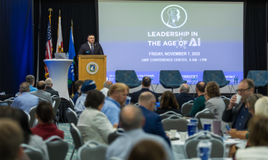 Person speaks at a podium in front of a seated audience. Projection reads, "Leadership in the Age of AI. Friday, November 7, 2025 UWF Conference Center, 9 a.m. - 1 p.m."