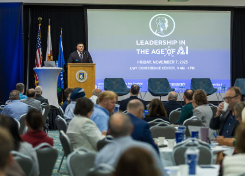 Person speaks at a podium in front of a seated audience. Projection reads, "Leadership in the Age of AI. Friday, November 7, 2025 UWF Conference Center, 9 a.m. - 1 p.m."