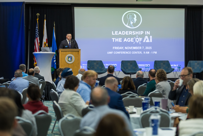 Person speaks at a podium in front of a seated audience. Projection reads, "Leadership in the Age of AI. Friday, November 7, 2025 UWF Conference Center, 9 a.m. - 1 p.m."