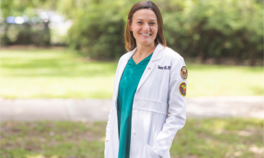 Faculty member in white medical coat smiles outdoors with hands in pockets.
