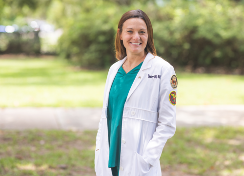 Faculty member in white medical coat smiles outdoors with hands in pockets.