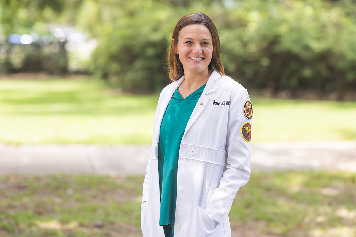 Faculty member in white medical coat smiles outdoors with hands in pockets.