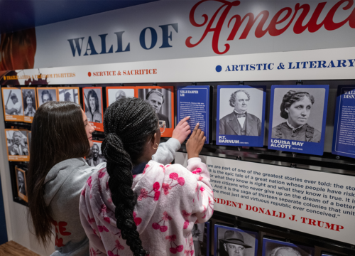 Two middle-school-aged children point at the mobile exhibit's Wall of America featuring photos of historical figures.