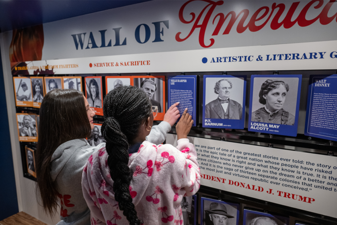 Two middle-school-aged children point at the mobile exhibit's Wall of America featuring photos of historical figures.